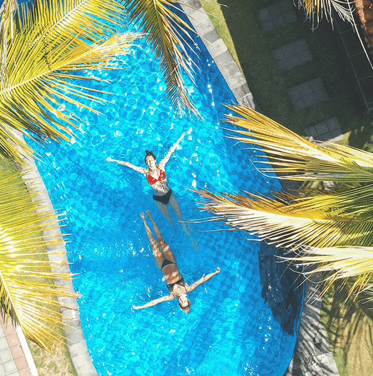 An aerial, close-up view of two women floating on their backs in a bright blue swimming pool, surrounded by the fronds of palm trees.