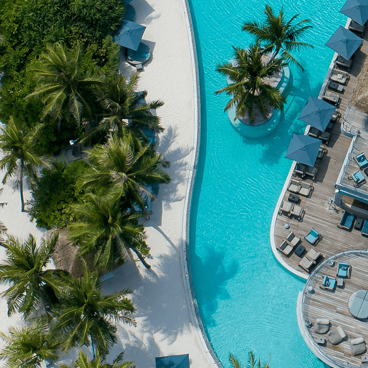An aerial view of a luxurious tropical resort pool area. The bright turquoise, curving pool is bordered by a white sand beach with palm trees and a wooden deck with lounge chairs and dark umbrellas.