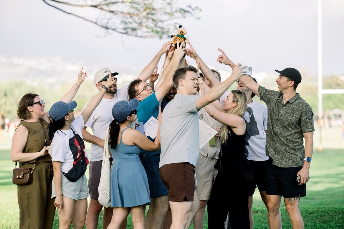 Image 9: A group of colleagues huddled together in a park, triumphantly lifting a small stuffed giraffe trophy into the air after winning a team competition.