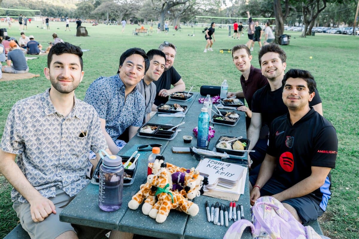 Image 10: Six coworkers gathered around a picnic table in a park, smiling with food and stuffed giraffe mascots on the table in front of them.