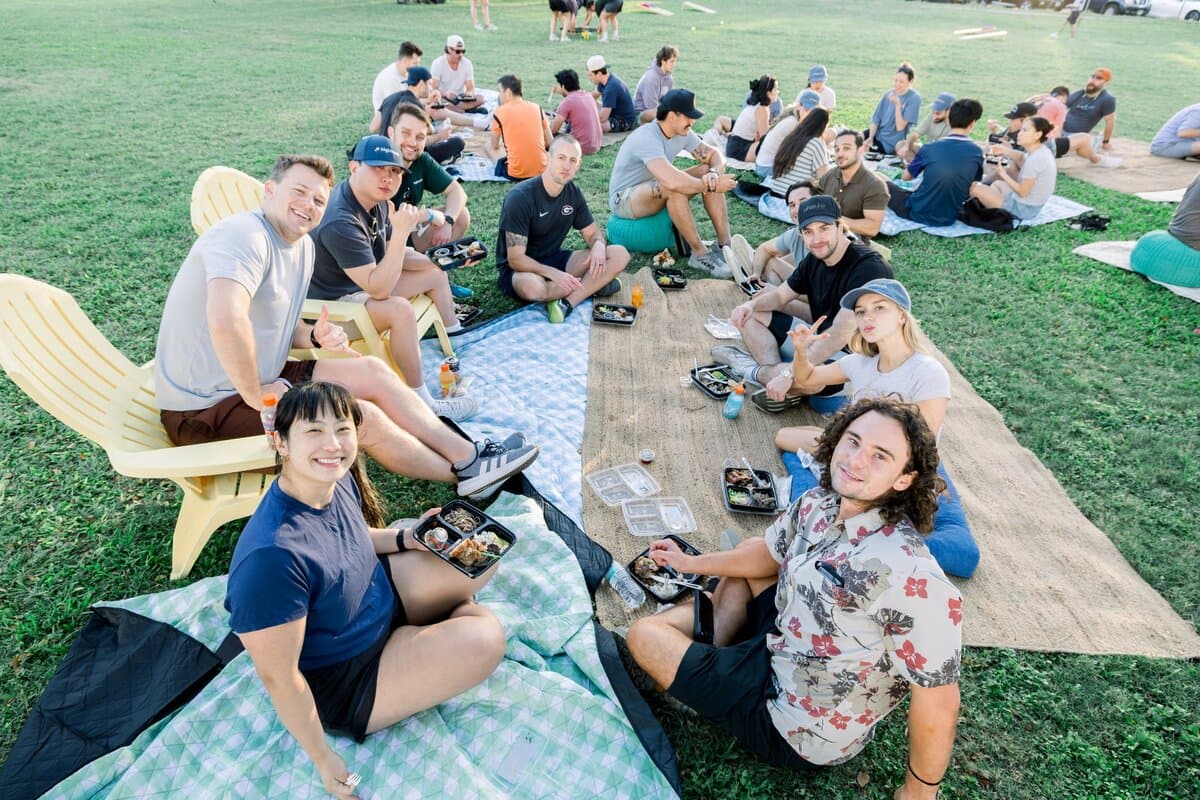 Image 11: A large group of employees eating and relaxing on blankets across a grassy park field at an outdoor team picnic.