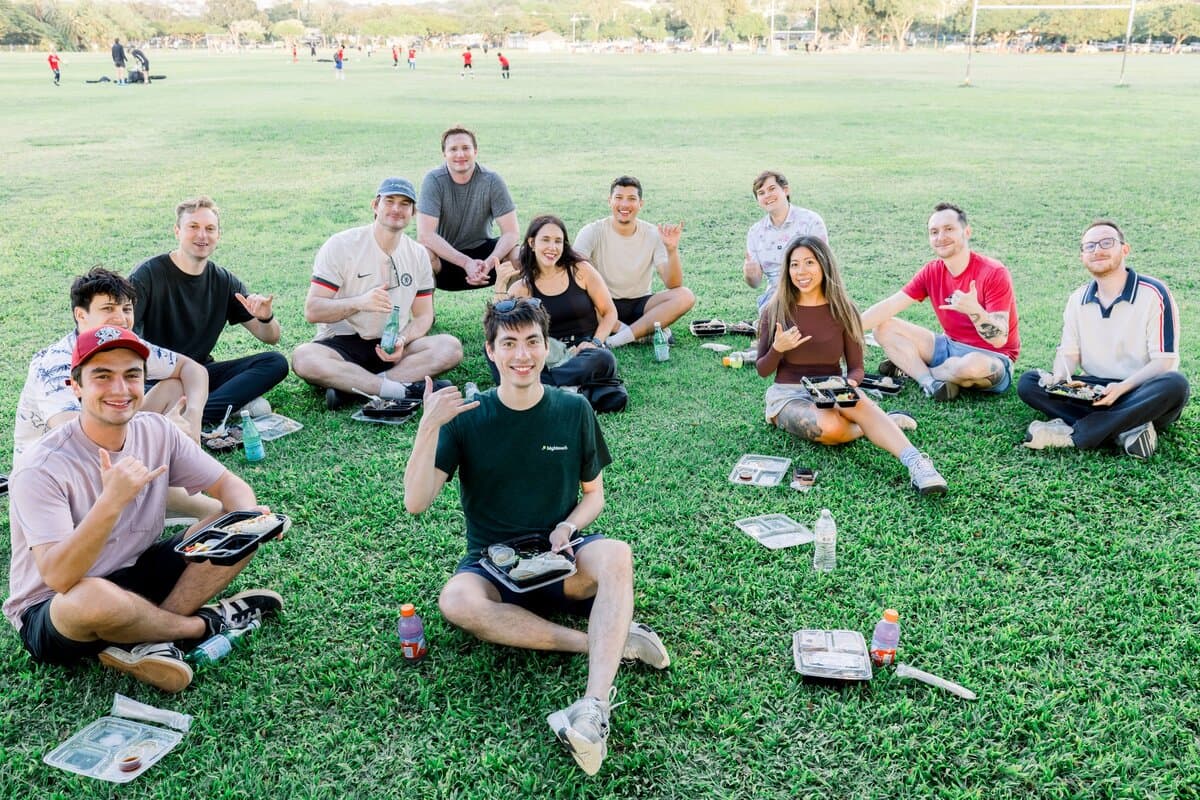 Image 12: About a dozen colleagues seated in a circle on the grass at a park, smiling and giving shaka signs at the camera.