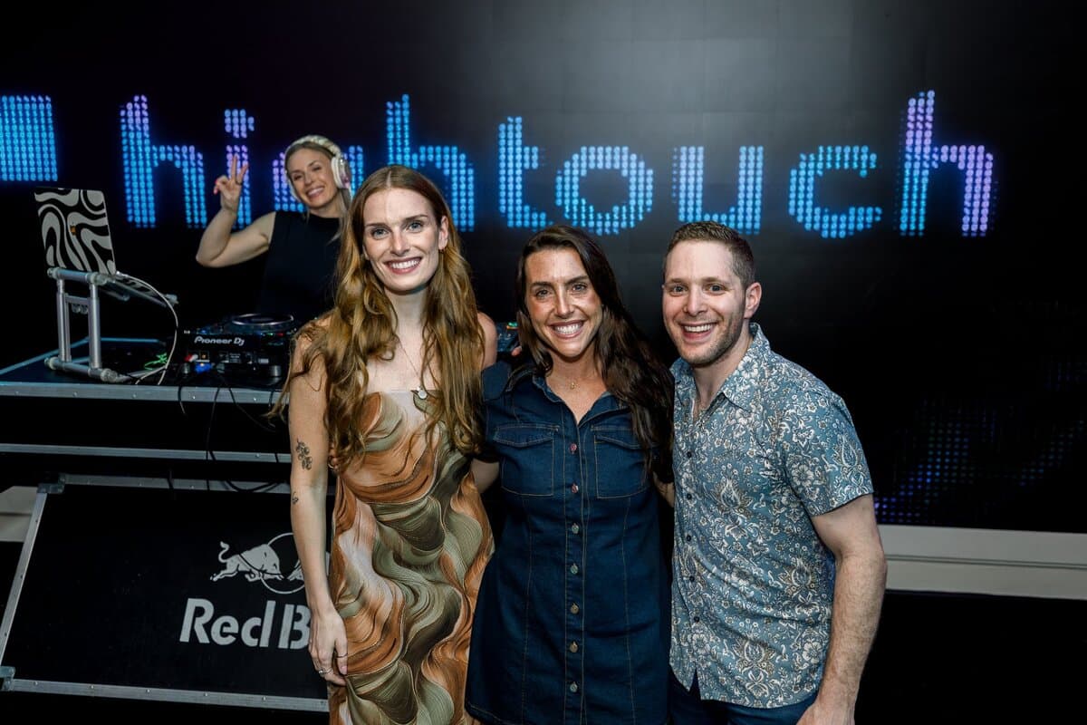 Image 13: Three people posing in front of a glowing "hightouch" LED sign with a DJ booth and Red Bull branding at a company evening party.