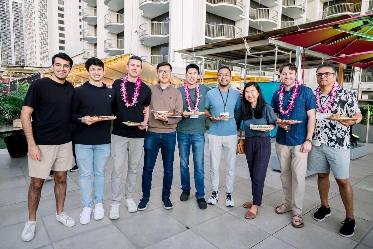 Image 3: Nine coworkers posing together outdoors, several wearing leis and holding plates of food at a company gathering.