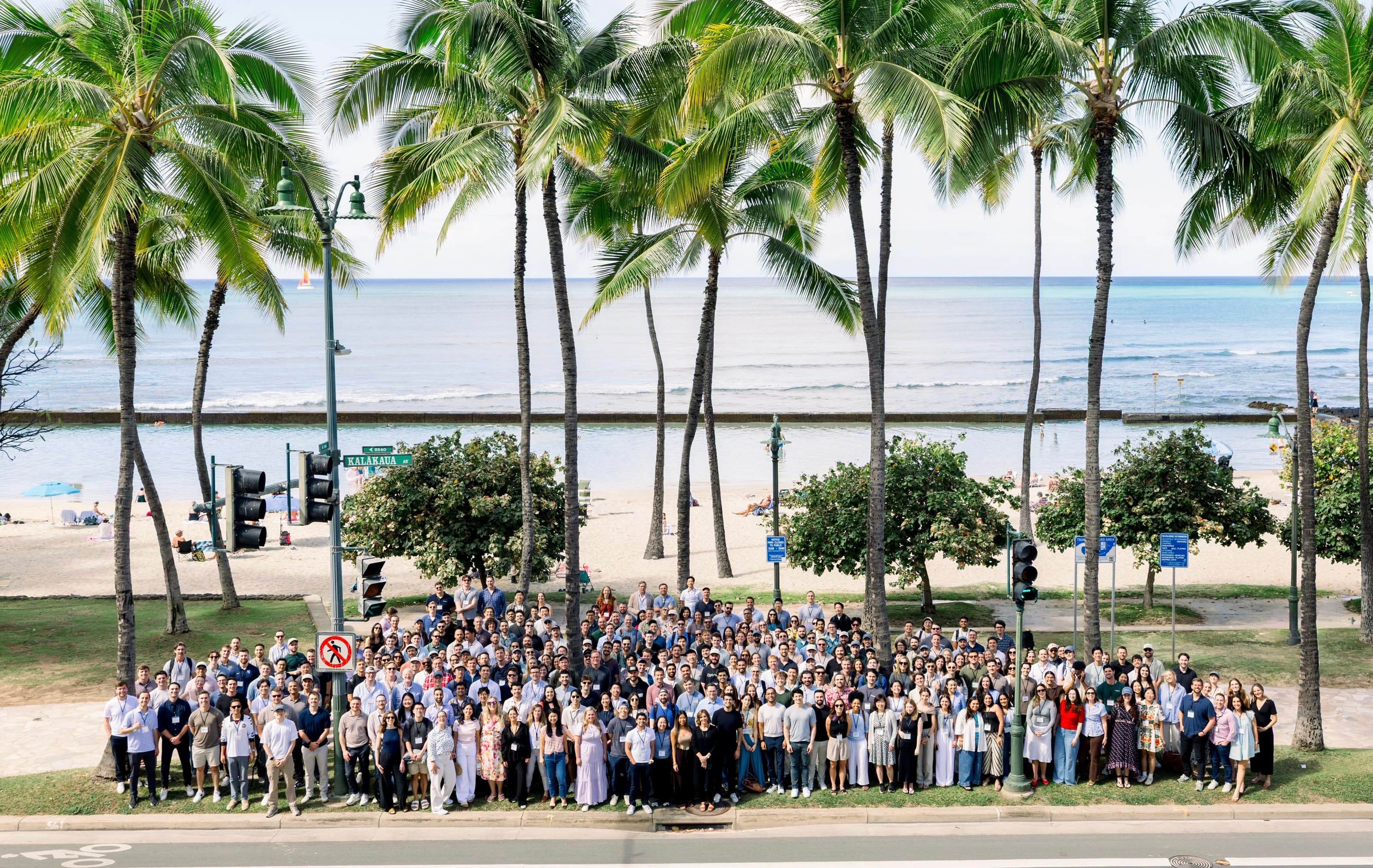Aerial view of a large group of several hundred employees posed together on the sidewalk along Waikiki Beach, with palm trees and the ocean behind them.