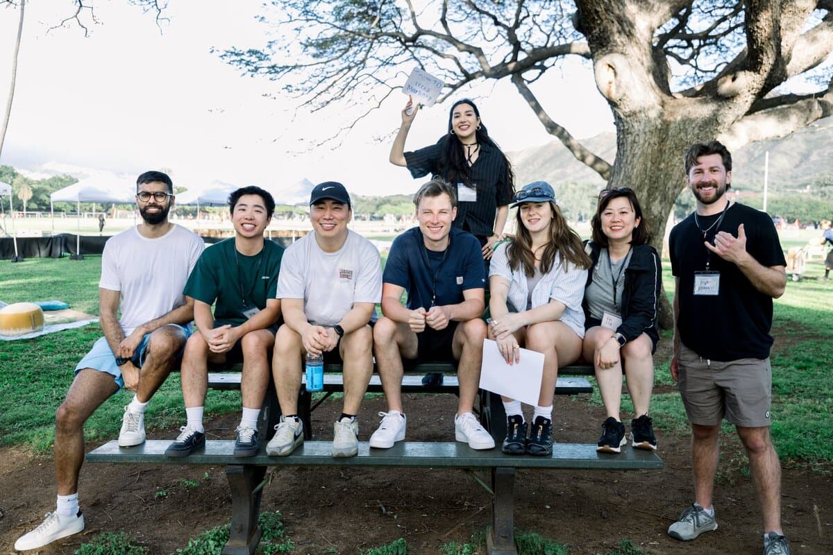 Image 8: A team of eight coworkers seated and standing around a picnic table at an outdoor park activity, one holding up a handwritten sign.