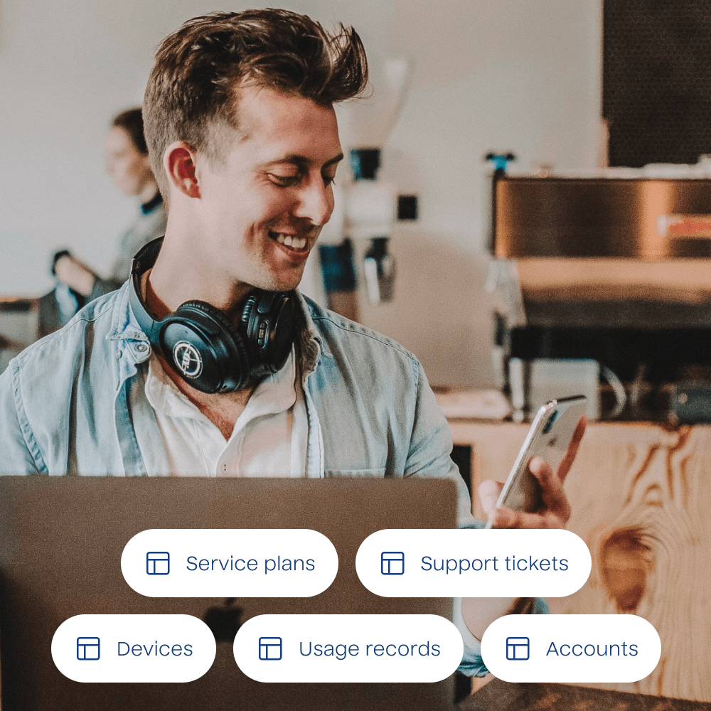 A photograph of a person holding a cell phone with a list of example data tables overlayed. The data tables included are for service plans, support tickets, devices, usage records, and accounts.